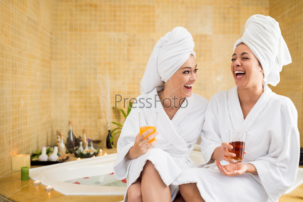 Cheerful women in bathrobes drinking juice: friends in spa salon
