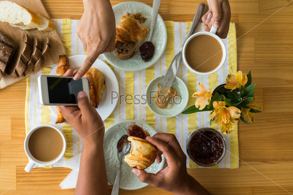 Hands of couple having breakfast and using smartphone, view from above