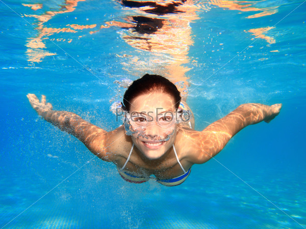 Woman swimming underwater in pool smiling. Young female\
swimmer with swim goggles at holiday resort.