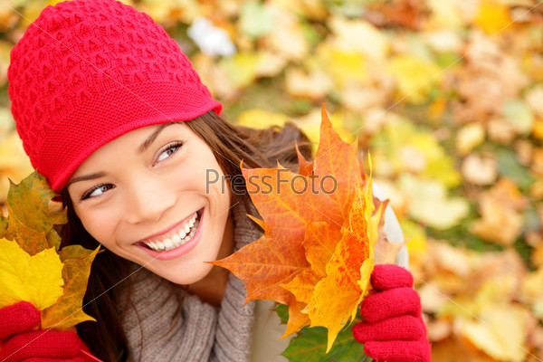 Fall woman looking at copy space holding fall leaves smiling happy and joyful. Lovely beautiful girl wearing red knit hat and gloves. Fall lifestyle concept image.
