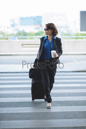 Stylish business lady with coffee and suitcase crossing the road