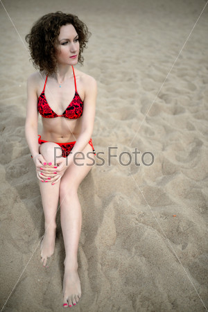 The beautiful girl with a curly hair in a red bikini sit on a beach