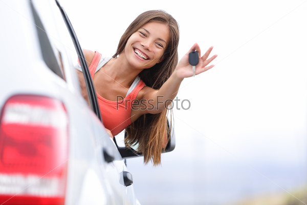 Car driver woman happy showing car keys out window. New car, rental or driving licence concept with young female model on road trip. Mixed race Asian Caucasian girl in her 20s.