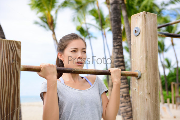 Fitness woman exercising on chin-up bar. Lady doing chin-ups training toned arms portrait outside on beach in summer.