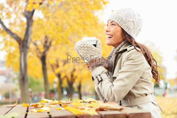 Fall concept - autumn woman drinking coffee on park bench under fall foliage. Beautiful young modern woman smiling happy and cheerful in trench coat.