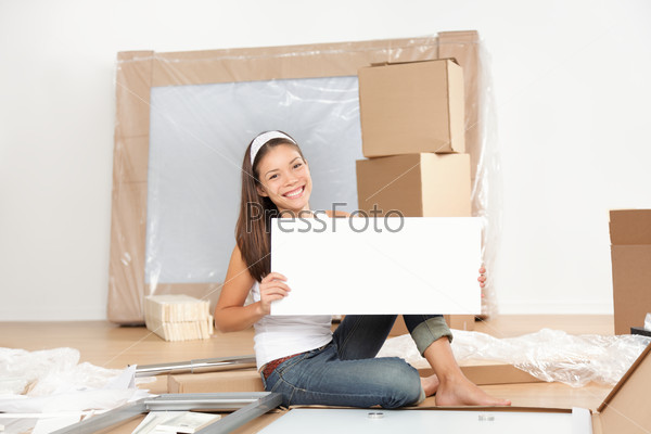 Moving sign woman. Woman moving in new home unpacking showing blank sign while doing furniture assembly of new table. Beautiful young mixed race Asian Caucasian young woman in new apartment condo