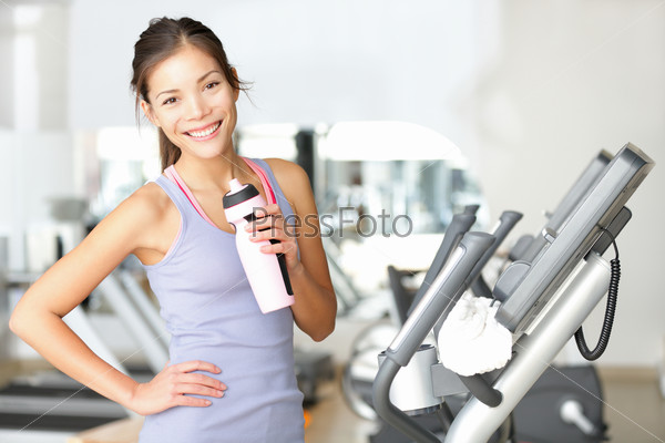 Gym woman working out drinking water smiling happy standing by moonwalker fitness machines. Beautiful fit young mixed race Caucasian / Chinese Asian female fitness model inside in fitness center.