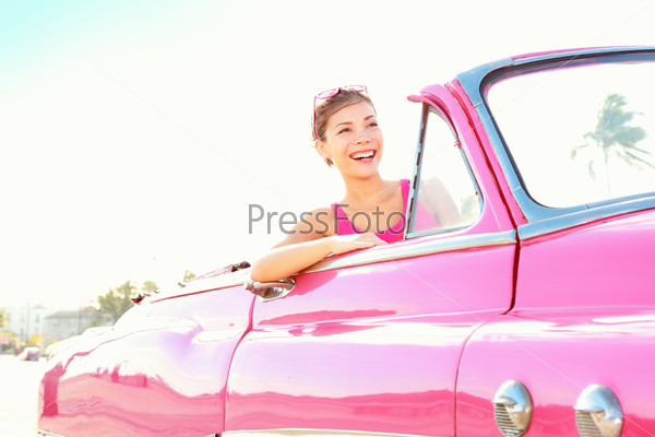 Retro woman smiling happy in old pink vintage car driving on road trip on beautiful summer day. Pretty multiracial Asian / Caucasian female model