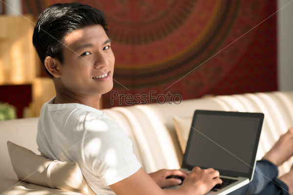 Cheerful Asian man sitting on the sofa and working on laptop
