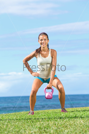 Fitness exercise woman lifting kettlebell during strength training exercising outdoors on grass by the ocean. Beautiful young fit fitness instructor doing one-arm kettlebell swing or snatch.
