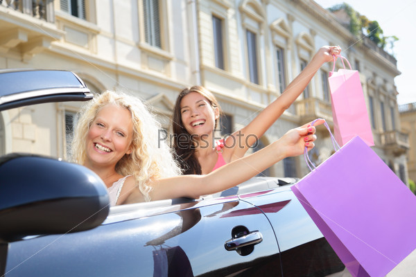 Car driver woman driving and shopping with girl friends holding shopping bags happy and excited on road trip travel vacation in convertible car in summer. Two beautiful women girlfriends shoppers.