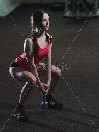 Young, beautiful, athletic girl performs squats with dumbbells in a gym. Hard light