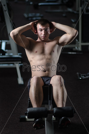 Young handsome man is training the abdominal muscles of the abdomen against the backdrop of the gym