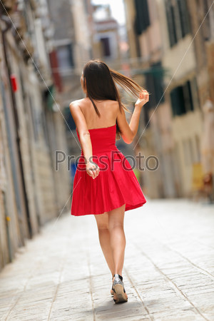 Woman in red dress walking in street in Venice, Italy cheerful and happy in rear view showing back of sundress. Pretty sexy brunette fashion model girl in her 20s.