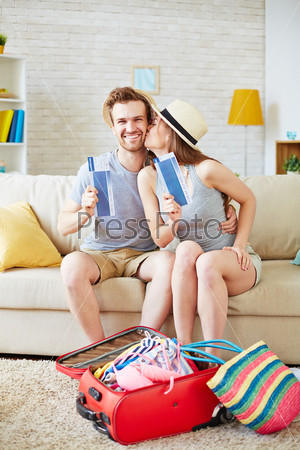 Happy couple with air tickets sitting on sofa with suitcase near by