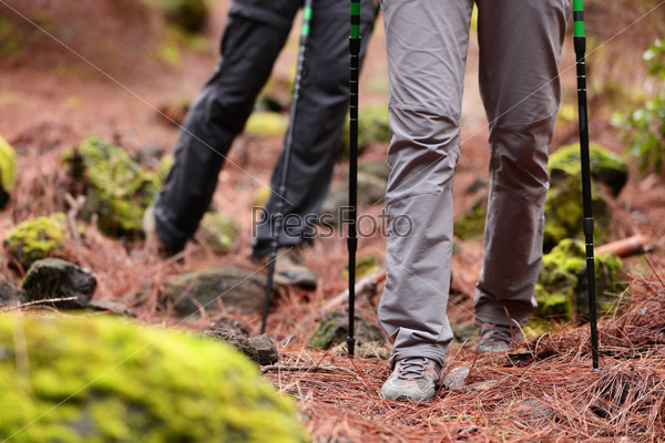 Hiking - Hikers walking in forest with hiking sticks on path trail in mountains. Close up of hiking shoes and boots. Man and woman hiking together.