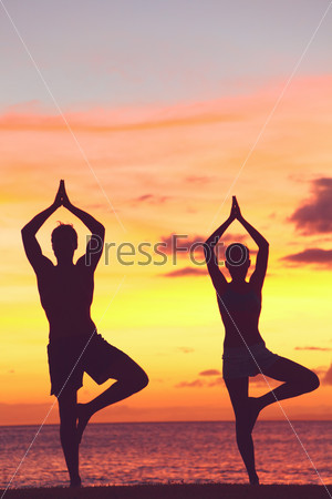 Yoga couple training in sunset in tree pose meditating outdoors by beach ocean sea. Man and woman working out training in serene ocean landscape. Silhouette against sunset