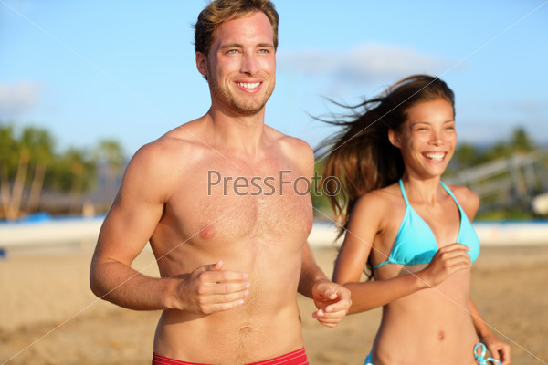 Running athletic couple jogging on beach training in beach wear swimsuit, bikini and shorts. Shirtless handsome man fitness model and beautiful mixed race asian caucasian woman happy and smiling.