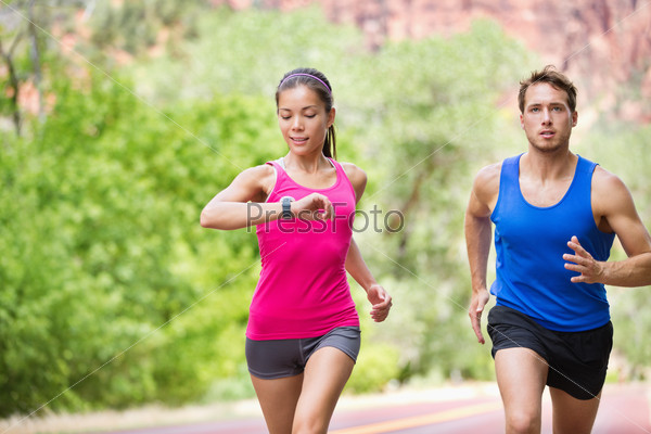 Runners - mixed multicultural couple training outside in\
nature. Fitness asian young smiling model checking time or pulse on\
heart rate monitor watch and male model jogging.