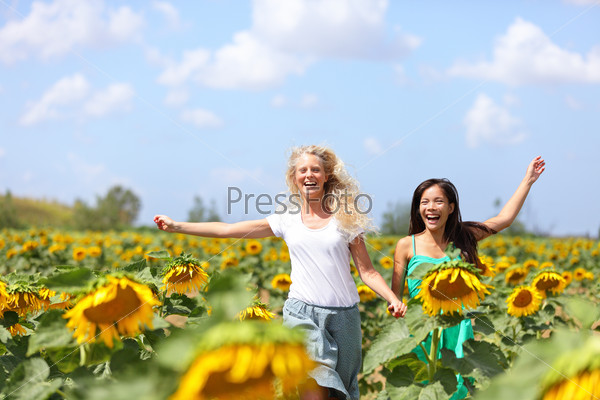 Two happy carefree young women running through field of sunflowers on a sunny summer day laughing and having fun with arms in the air. Beautiful young multiracial girlfriends, Asian and Caucasian girl