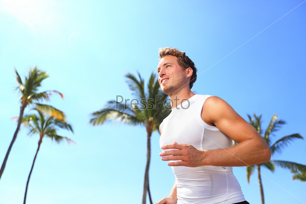 Sport man running. Male athlete runner jogging in compression\
t-shirt top training on palm trees beach. Fit handsome male fitness\
model jogging alone training for marathon run. Man in his\
twenties.