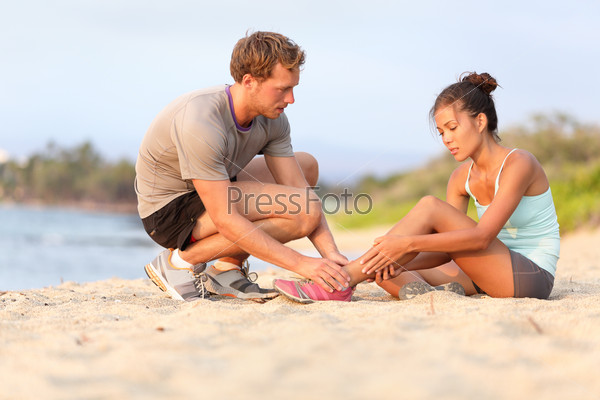 Injury - sports woman with twisted sprained ankle. Asian fitness female model sitting on beach sand getting help from Caucasian male touching her ankle.