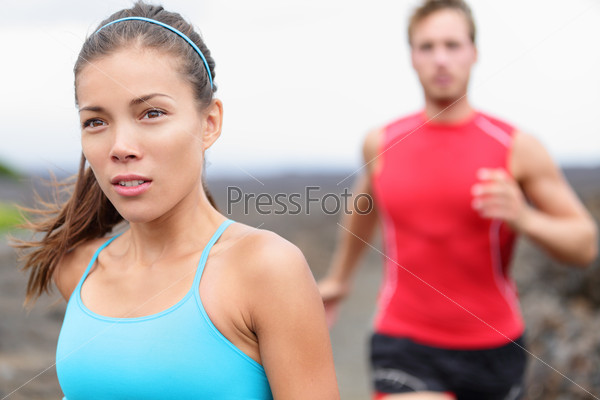 Woman runner closeup - running couple. Close up of girl jogging outside with man in background. Fit athlete couple training together outdoors. Mixed race Asian Caucasian female fitness model.