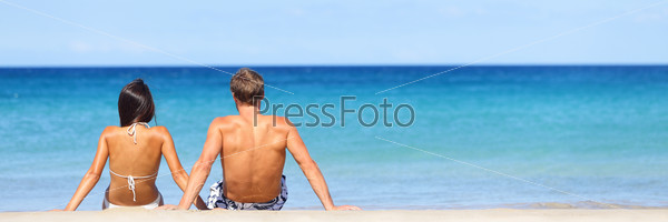 Beach travel banner - romantic couple relaxing on vacation enjoying ocean view together sitting in the sand embracing and hugging. Beautiful young multiracial couple, Asian woman, Caucasian man.
