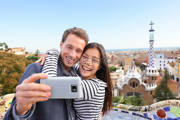 Travel couple happy talking selfie self-portrait with smartphone in Park Guell, Barcelona, Spain. Beautiful young multiracial couple looking at camera taking photo with smart phone smiling in love.