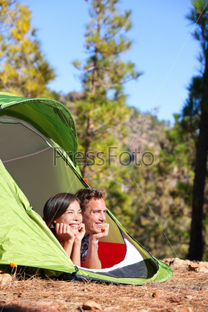 Camping couple in tent romantic looking at view in forest. Campers smiling happy outdoors in forest. Multiracial couple having fun relaxing after outdoor activity. Asian woman, Caucasian man.