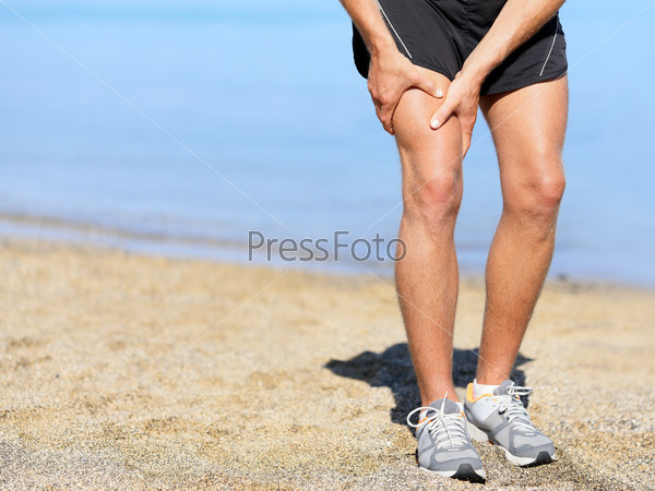 Muscle injury. Runner man with sprain thigh muscle. Athlete\
in sports shorts clutching his thigh muscles after pulling or\
straining them while jogging on the beach wearing running\
shoes.
