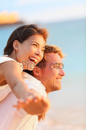 Couple on beach having fun laughing in love on romantic honeymoon travel vacation summer holidays romance. Young happy lovers, Asian woman and Caucasian man doing joyful piggybacking ride outdoors.
