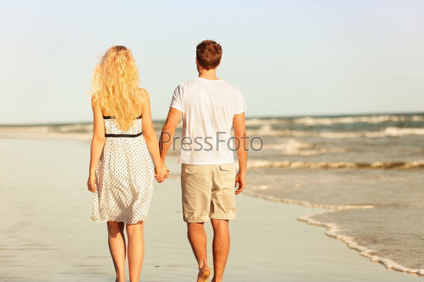 Beach couple holding hands walking at sunset by ocean sea. Beautiful casual young lovers hand in hand walking away with back at rear view. Summer vacation beach travel holidays concept.