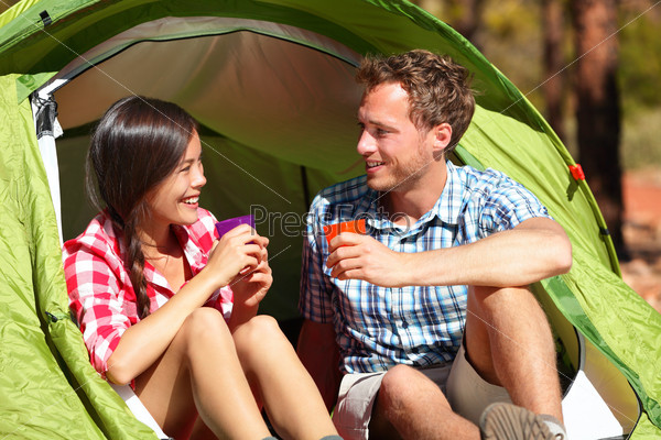 Camping couple drinking water in tent smiling happy outdoors in forest enjoying sun at looking at view. Happy multiracial couple relaxing after outdoor activity hiking. Asian woman, Caucasian man.