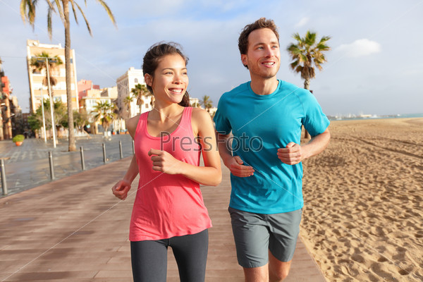 Running couple jogging on Barcelona Beach, Barceloneta. Healthy lifestyle people runners training outside on boardwalk. Multiracial couple, Asian woman, Caucasian fitness man working out, Spain.
