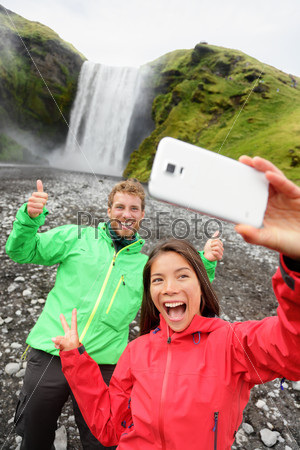 Selfie couple taking funny selfie smartphone picture of waterfall outdoors in front of Skogafoss on Iceland. Couple visiting famous tourist attractions and landmarks in Icelandic nature landscape.