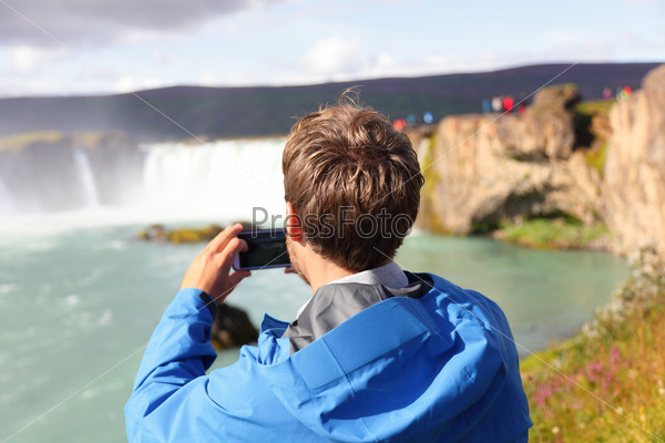Tourist taking photo with smartphone of waterfall Godafoss on Iceland. Man taking picture with smart phone camera on travel visiting tourist attractions and landmarks in Icelandic nature on Ring Road.