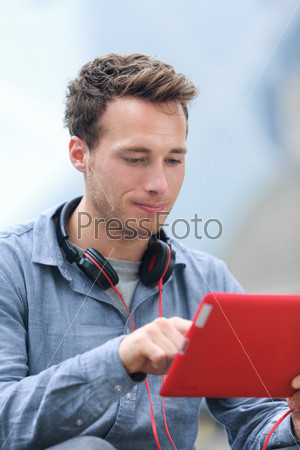 Urban young professional man using tablet computer sitting outside using app on 4g wireless device wearing headphones. Casual young urban professional male in his late 20s.