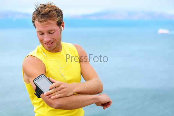Running training music on smartphone app. Runner man listening to music adjusting settings on armband for smart phone. Fit male fitness model working outdoor by water.