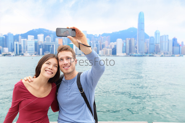 Selfie tourists couple taking self-portrait picture photos in Hong Kong enjoying sightseeing on Tsim Sha Tsui Promenade and Avenue of Stars in Victoria Harbour, Kowloon, Hong Kong. Travel concept.
