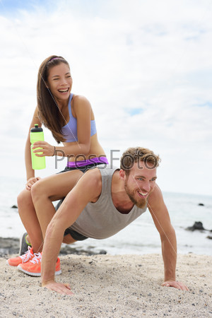 Fitness couple training doing funny push-up on beach during workout. Woman playful having fun sitting on boyfriend to test his strength with heavy weight.