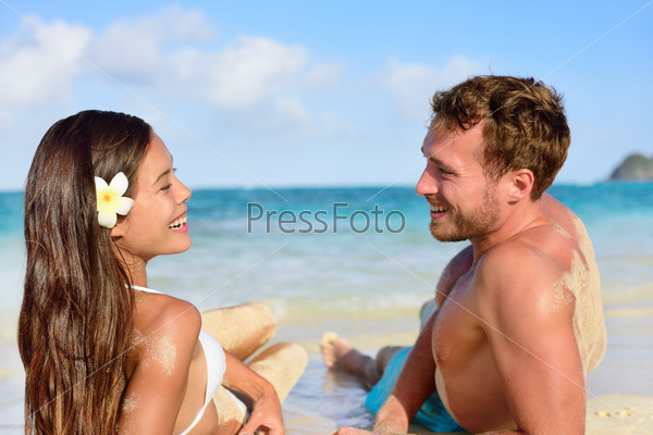 Vacation couple relaxing on beach tanning laughing talking together. Portrait of two young adults mixed race asian caucasian having fun during travel holidays.