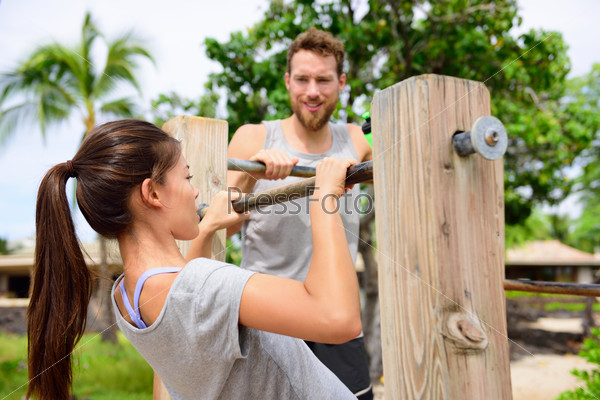 Fitness couple training on chin-up bar together. Woman helped by friend or personal trainer in coaching session supervising exercises on outdoor beach gym.