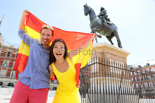 Madrid people showing Spain flag on Plaza Mayor cheerful and happy in Spain. Cheering celebrating young woman and man holding and showing flags to camera on the famous square in front of statue.