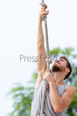 Fit strong man cross training climbing rope. Male adult exercising rope climb as part of crossfit workout routine outdoors on beach gym.
