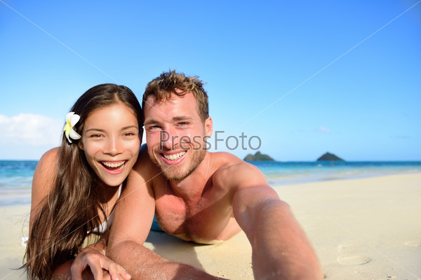 Couple relaxing on beach taking selfie picture with camera smartphone. Young multiracial couple on getaway vacation in Hawaii lying down looking at camera. Candid closeup angle looking candid real.