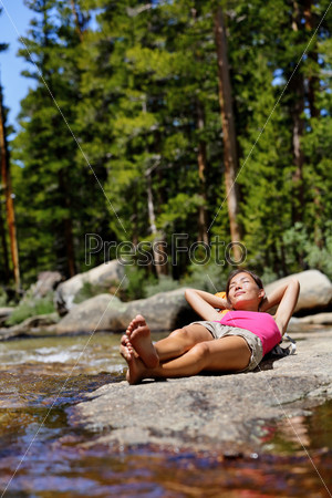 Hiking girl relaxing sleeping in nature next to river creek. Tired hiker resting lying down outdoors taking a break from hike. Young Asian woman in forest in Yosemite national park, California, USA.