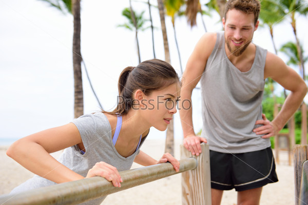 Fitness instructor coaching and helping woman doing push-ups on cross fit horizontal bar station on beach. Arm press pushups easy exercises.