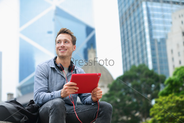 Urban man using tablet computer sitting in Hong Kong outside using app on 4g wireless device wearing headphones. Casual young urban professional male in his late 20s. Hong Kong Central.
