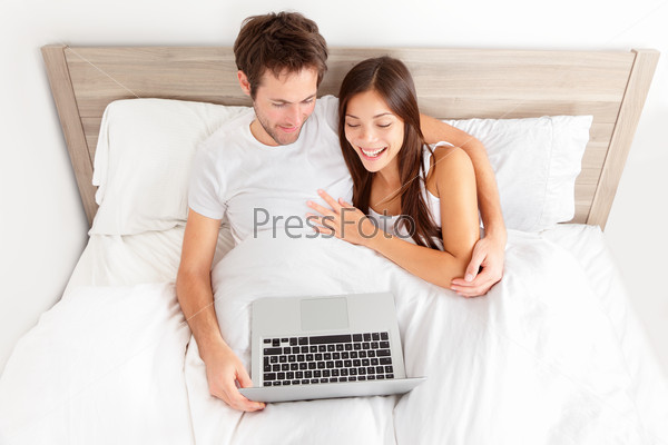 Young couple relaxing in bed. Happy affectionate young couple relaxing in bed enjoying themselves laughing in amusement with their laptop computer. Interracial couple, Asian woman, Caucasian man..